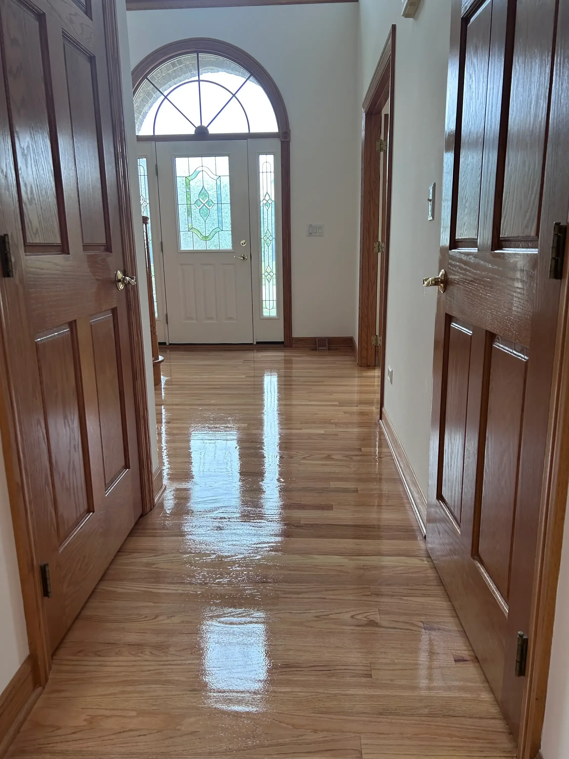 Hardwood foyer flooring with glossy finish, dark wood doors, and arched stained glass entry